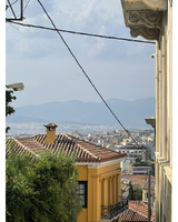 A view of rooftops and trees with a crowded city and mountains in the background. The sky is hazy and clouded with power lines running across the frame.