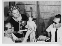 Three people work on a sculpture; one holds a photo of a suited man. Books including "THE LYNDON JOHNSON STORY" lie nearby.