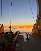 Ship deck at sunset with ocean and ice visible, featuring cables and equipment.