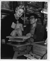 Two individuals work on a small clay sculpture in a cluttered room.