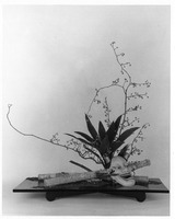 Ikebana arrangement with branches, leaves, a mask, and logs on a flat base.