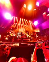 Orange and pink lights shine on a stage with musicians and equipment. Audience members hold cell phones and cameras up to capture the concert. Behind the raised drum kit, a segmented projection screen displays an image.