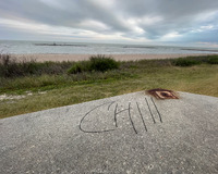 A cement platform rests on a grassy shoreline. The cement is in the foreground with one corner visible, stretching towards the water. Grass and low scrub can be seen between the cement and the sandy shore.  Three break walls or other pier like structures are visible in the water running parallel with the coast. The sky is overcast and the tone is generally grey. A discolored mark at the corner of the cement looks like rust or the remainder of a small fire. The word "Chill" is written in large black letters across the cement.