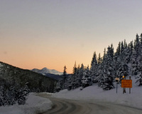 A snowy mountain road at dusk with snow-covered trees and a gradient sky.