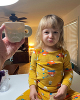A child in yellow vehicle-themed pajamas sits on a countertop next to a hand holding a mug with a sculpted face.