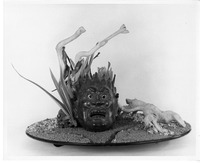 Artistic arrangement with a fierce mask, twisted structures, and natural elements on a sandy base.