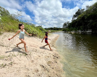 A bright clouded sky is visible above a river. The near shore is steep and sandy with vegetation further up the rise. The far shore is rocky and covered in trees. Two young swimmers excitedly approach the water, smiling in their swimsuits.