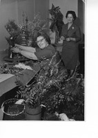 Two women arranging flowers and a small figurine on a table, surrounded by foliage and decorative elements.