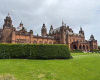 A historical building with reddish-brown stone, turrets, and ornate architecture, set behind a green lawn and hedge under an overcast sky.