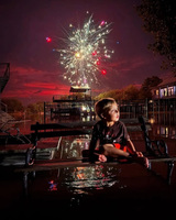 A child sitting on a bench watching fireworks at night with reflections on wet ground.