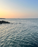 Sunset over a calm sea with rippling waves, silhouetted rocks, and a distant pier.