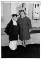 Two individuals in traditional and modern clothing stand in front of art-adorned walls.