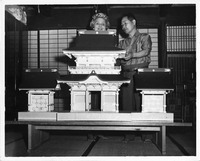 Two people look at a model of a traditional Japanese building with detailed roofs and panels on a table. Shoji screens are in the background.