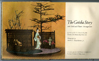 Cover of 'The Geisha Story' featuring a traditional Japanese doll in a kimono posed beside a flower arrangement with trees, lantern, and bamboo fence.