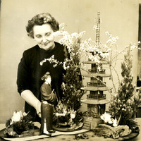 Older woman in a dark dress stands behind a detailed miniature Japanese scene with a figurine, trees, plants, and a pagoda.