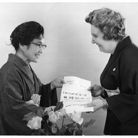 Two women in kimonos smiling at each other while holding a paper. A bouquet of roses is in the foreground.