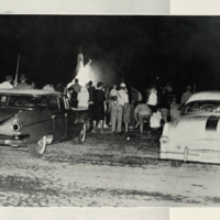 Students outside at a bonfire. They are surrounded by a TP fire with two cars behind them. TheSilverKing1959. There is two old school cars and a large fire. 