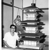 Two people beside a model of a multi-tiered pagoda indoors.