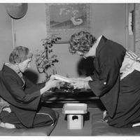Two women in kimonos engage in a traditional Japanese tea ceremony, with a bonsai on a table between them.