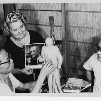 Three people work on a sculpture; one holds a photo of a suited man. Books including "THE LYNDON JOHNSON STORY" lie nearby.