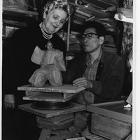 Two individuals work on a small clay sculpture in a cluttered room.