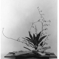 Ikebana arrangement with branches, leaves, a mask, and logs on a flat base.