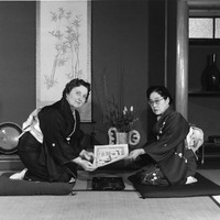 Two women in kimonos exchange a document while seated on cushions in a traditional Japanese room.
