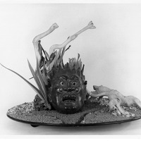 Artistic arrangement with a fierce mask, twisted structures, and natural elements on a sandy base.