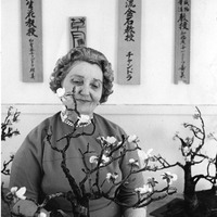 Woman holding a flower arrangement with Japanese calligraphy plaques on the wall behind her.