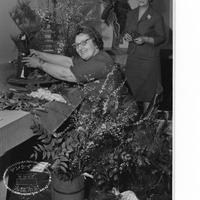 Two women arranging flowers and a small figurine on a table, surrounded by foliage and decorative elements.