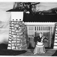 Performer in traditional costume stands by a Japanese-style gate with mountains in the background.