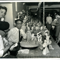 People working with and observing small sculptures in a workshop setting; sculptures and figures cover the tables.