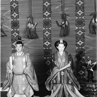 Two traditional Japanese dolls in front of a patterned backdrop with tassels.