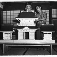 Two people look at a model of a traditional Japanese building with detailed roofs and panels on a table. Shoji screens are in the background.