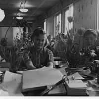 Students seated in a classroom with floral arrangements on desks; papers and camera in the foreground.