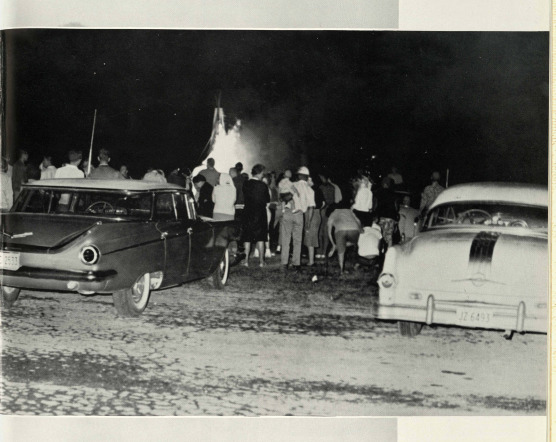 Students outside at a bonfire. They are surrounded by a TP fire with two cars behind them. TheSilverKing1959. There is two old school cars and a large fire. 